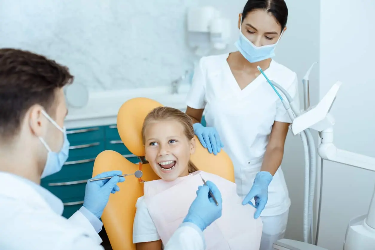 A child smiling in a dentist chair next to a sitting pediatric dentist and standing hygienist