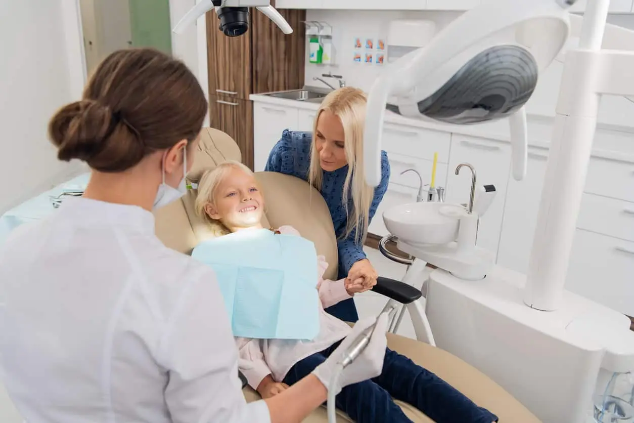 A child smiling in a dentist chair next to her pediatric dentist and mom